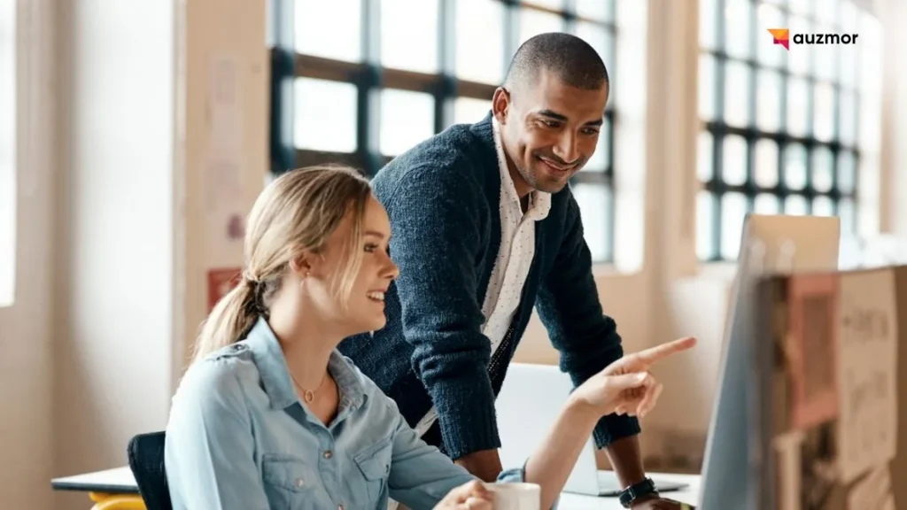 Mentor pointing at a screen while guiding a smiling employee at her desk
